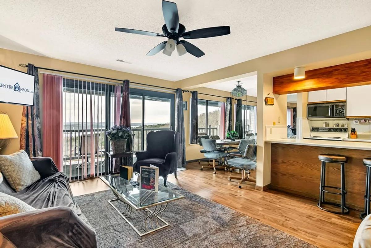 A warm, inviting interior scene featuring a sofa, coffee table, dining area, large windows letting in natural light, and a kitchen bar. The layout supports social gatherings and relaxation, with views extending out to the balcony and surrounding landscape.