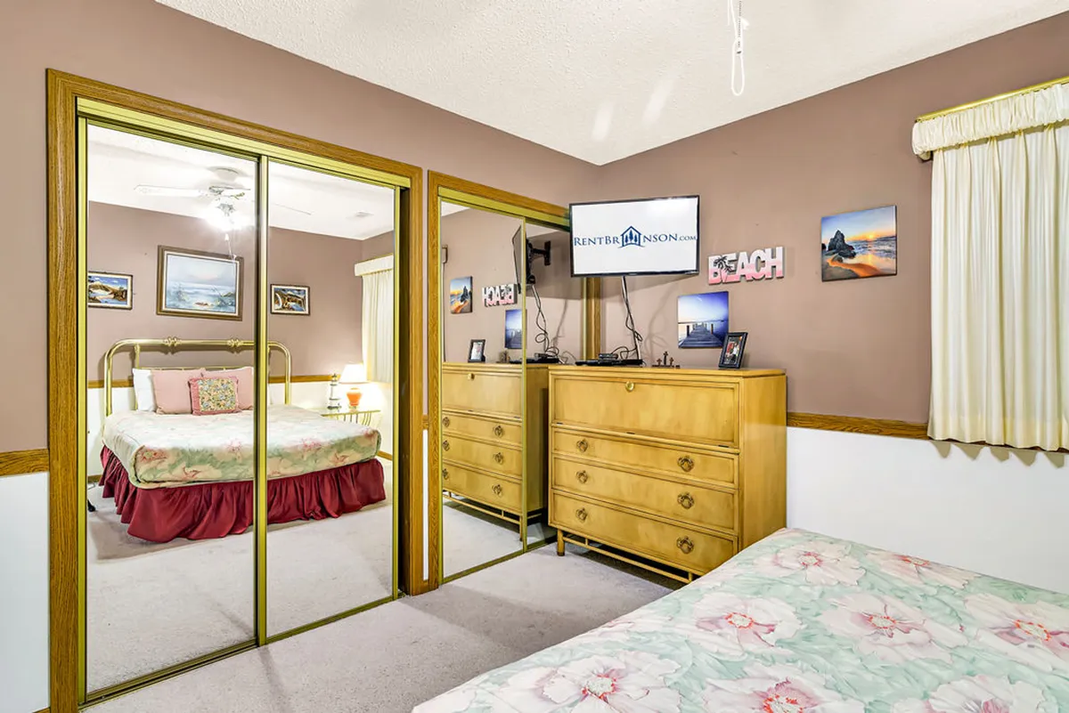 A cozy bedroom with floral bedding on a comfortable bed, a large mirrored closet, wooden dresser, and a TV mounted on the wall. Light streams in, giving the space a warm, welcoming feel.