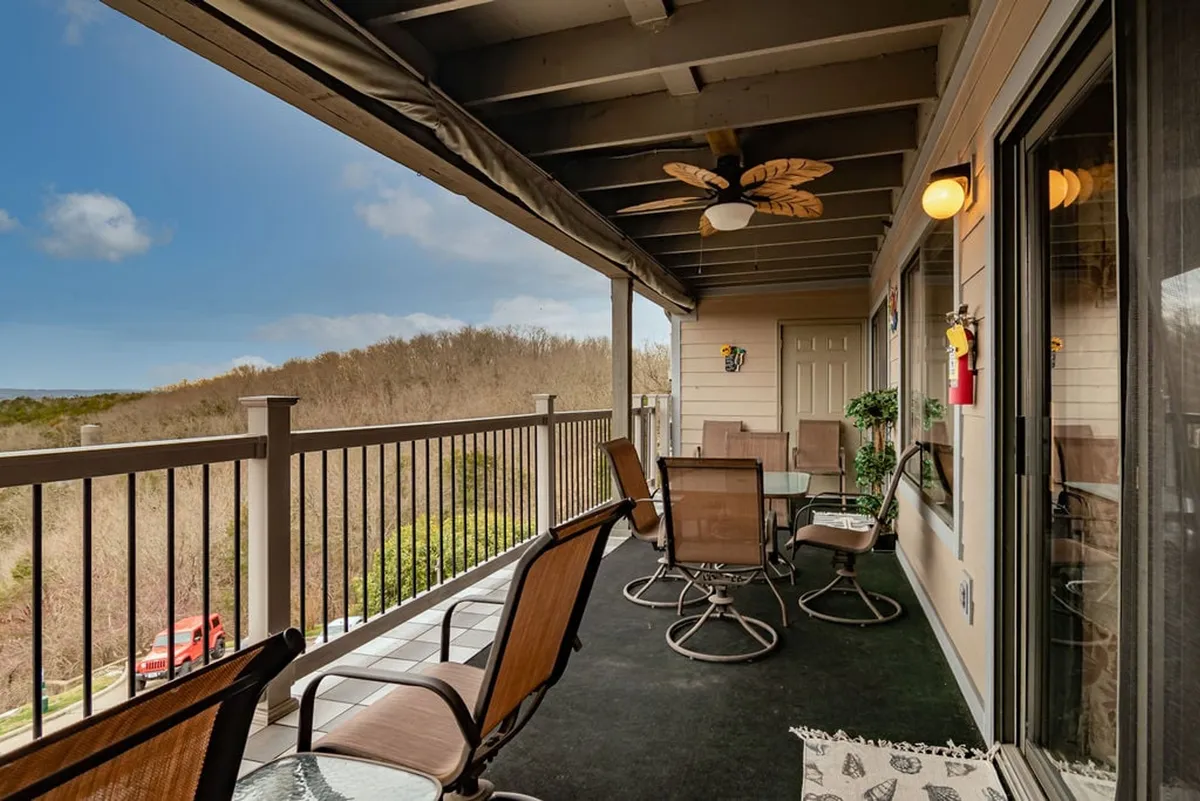 A serene exterior balcony with a glass-topped table and comfortable seating, offering a sweeping view of the rolling hills and natural scenery. The ceiling fan hints at comfort for long afternoons outside, while the open-air setting connects directly to the landscape of Table Rock Lake.