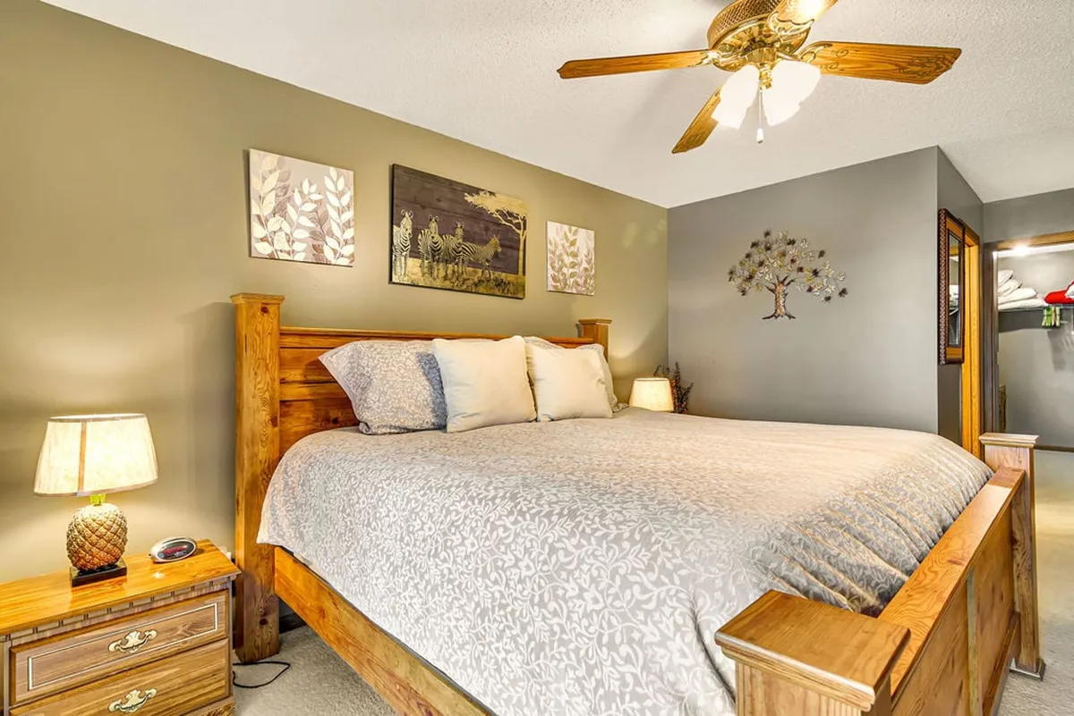 A spacious bedroom featuring a king-size bed with crisp white linens, a wooden nightstand, ceiling fan, and decorative wall art above the headboard. Soft sunlight filters into the room, creating an inviting and restful atmosphere.