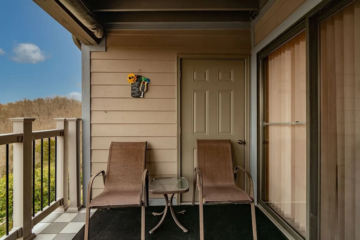 A cozy exterior patio setting featuring two comfortable chairs and a small glass table, set against a backdrop of decorative wall art. Soft morning light filters through nearby trees, creating a peaceful and relaxed atmosphere ideal for enjoying morning coffee or watching wildlife. The space feels accessible, inviting, and directly connected to the outdoors.