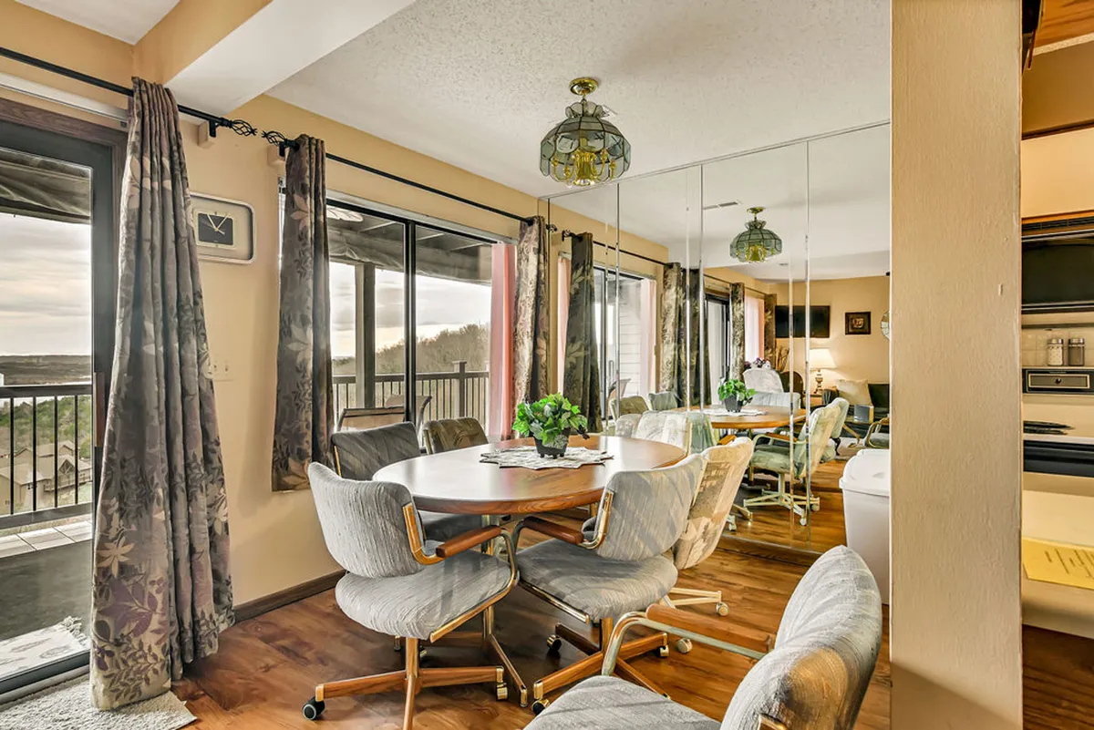 A cozy dining area featuring a substantial dining table, large windows letting in natural light, a decorative lamp, and a mirrored wall that enhances the room’s spacious feel. The scene suggests a warm, communal space ideal for family meals or gathering with friends.